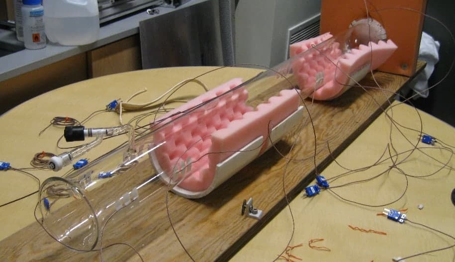 A long, clear, thin-walled glass cylinder lying on a bench, cradled in pink foam, with a valve on a side arm and thermocouple wires attached along its length.